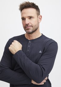 Mørkeblå langermet henley-skjorte med tre-knapps placket, ribbet tekstur og trekantet logo-detalj på brystet.