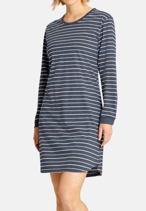 Frau trägt ein marineblaues, langärmeliges Kleid mit horizontalen weißen und hellblauen Streifen, steht vor einem schlichten weißen Hintergrund.