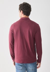 Langermet, burgunderfarget polo skjorte med en myk tekstur og klassisk krage, kombinert med lyseblå jeans. Navellengde og tettsittende design.