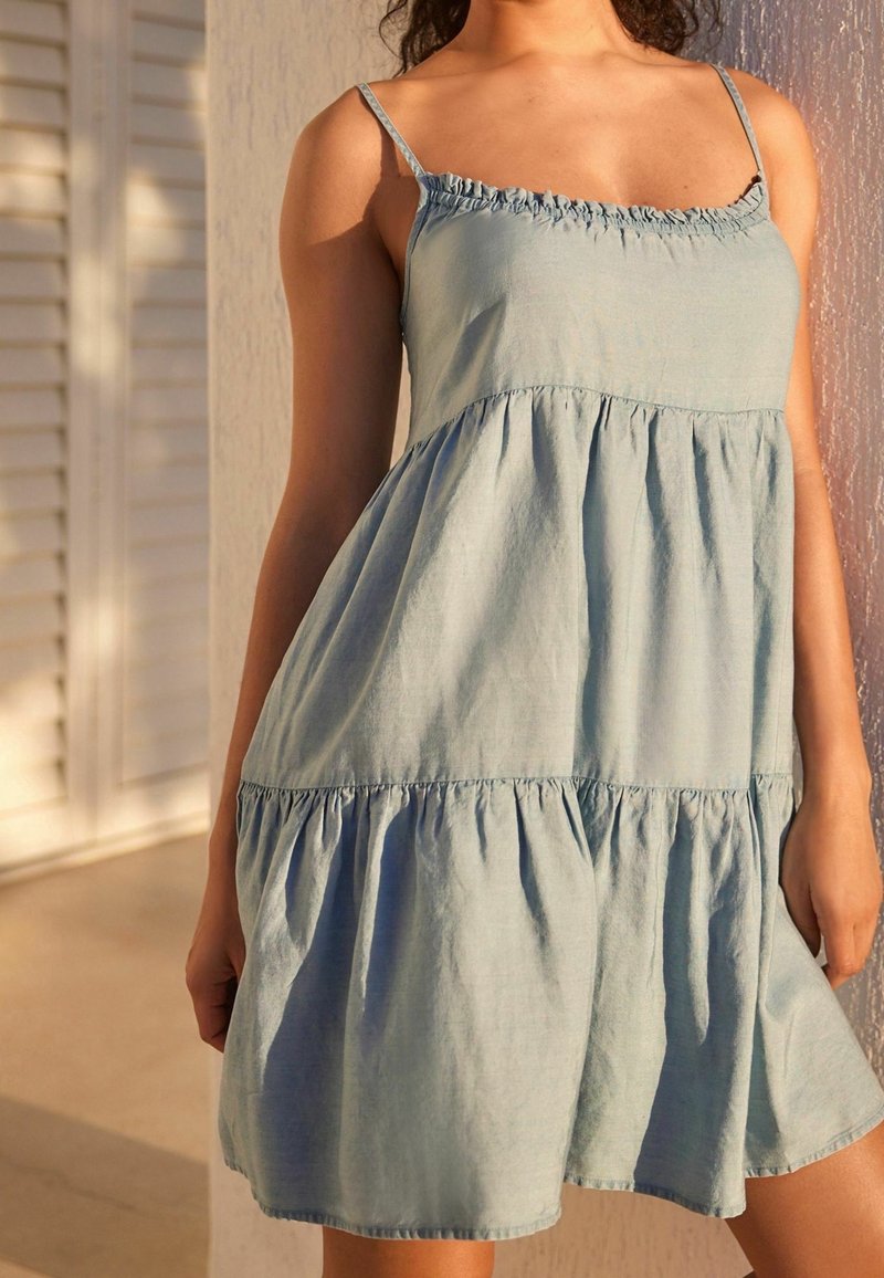 Femme portant une robe légère bleue ciel sans manches à étages avec de fines bretelles, debout contre un mur texturé sous une lumière douce.