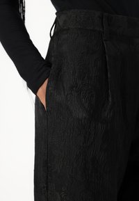 Pantalons noirs à motif avec un tissu texturé, présentant une taille haute et des poches latérales, montrés de côté avec une main dans une poche.