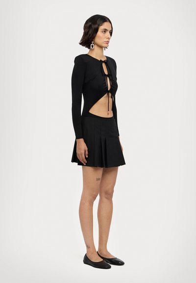 Robe noire à manches longues avec découpes et un détail de nœud à l'avant, présentant une jupe plissée et une texture lisse. Portée avec des ballerines noires.