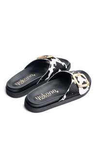 Black and white cow print mules with a wide strap, gold buckle detail, cushioned insole, and flat black rubber outsole.