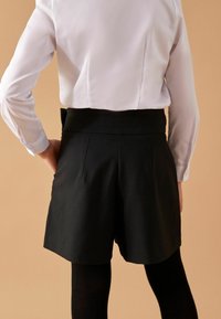 Visoko pasu črne kratke hlače iz teksturirane tkanine, z nabori na hrbtni strani in žepi, v kombinaciji s svetlo obarvano srajco in črnimi leggins.