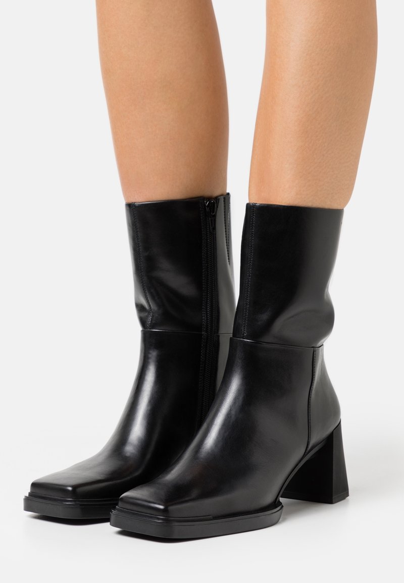 Bottes en cuir noires mi-mollet avec bouts carrés, talons blocs et fermetures éclair latérales portées sur des jambes nues sur fond blanc.