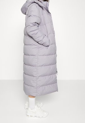 Osoba nosící dlouhý světle šedý péřový kabát s rukama v kapsách, bílé ponožky a robustní bílé tenisky na jednobarevném bílém pozadí.