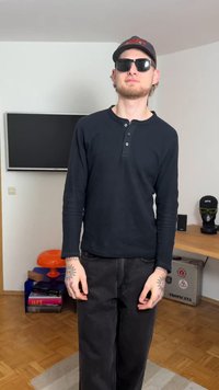 Black long-sleeve henley shirt with buttons, black trousers, sunglasses, and cap. Notable tattoos on the arms. Light-colored flooring and furniture.