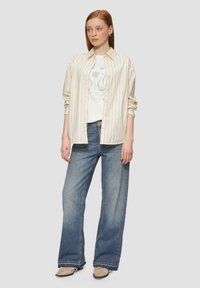 Gestreept overhemd in crème en lichtbeige, gedragen over een graphic t-shirt, gecombineerd met een wijde blauwe jeans en beige schoenen.