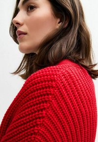 Röd stickad tröja med en strukturerad, ribbad design. Närbild från profilsidan som visar plaggets form och material.