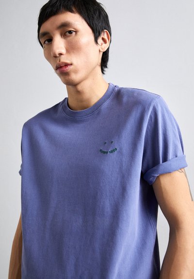 Μωβ βαμβακερό μπλουζάκι με στρογγυλή λαιμόκοψη, γυρισμένα μανίκια και πράσινη κεντητή λεπτομέρεια με χαμογελαστό πρόσωπο στο στήθος.