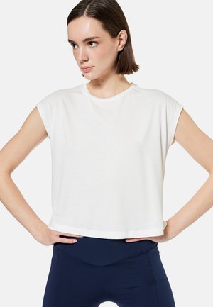 Jonge vrouw met kort bruin haar, draagt een los wit mouwloos topje en marineblauwe legging, handen op de heupen, kijkt opzij.