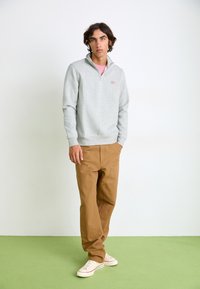 Lys grå zip-up sweatshirt med høj krave, båret over en lyserød skjorte. Tan bukser med synlige syninger og lommer. Hvide sneakers.