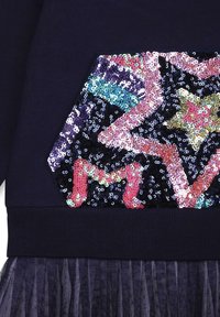 Mörk tyg med en flerfärgad paljettapplikation som har en stjärna och bokstaven "M", ovanför en mörk plisserad tylskjol.