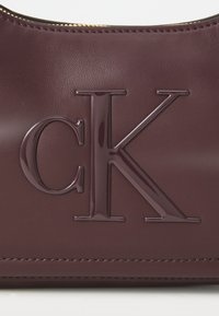 Bordeaux lederen handtas met een glanzend reliëf "CK" logo, een soepele textuur, ritssluiting en fijne stiksels langs de randen.