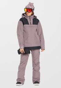Conjunto de snowboard para exteriores que incluye una chaqueta impermeable morada y negra con un bolsillo frontal, combinada con pantalones de nieve a juego y gafas de protección.