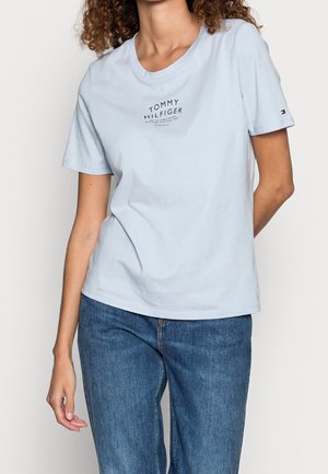 Kobieta ubrana w jasnoniebieską koszulkę Tommy Hilfiger z logo na piersi oraz niebieskie dżinsy, stojąca z jedną ręką za plecami.