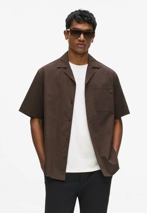 Férfi sötétbarna rövid ujjú gombos ingben, fehér póló fölött, fekete nadrágban és nagy sötét napszemüvegben, kezével a zsebében, semleges háttér.