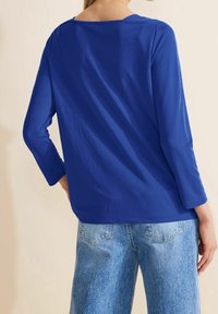 Maglia blu a maniche lunghe con scollatura ampia, realizzata in tessuto morbido, abbinata a jeans in denim blu chiaro con tasche posteriori.