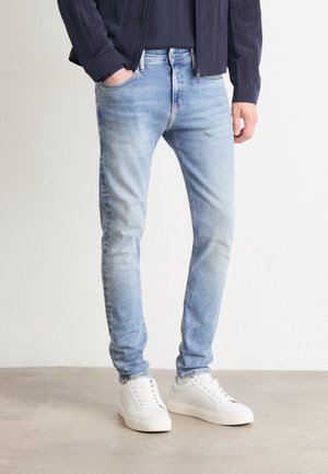 Mężczyzna ubrany w jasnoniebieskie dopasowane jeansy, czarną bluzkę i białe trampki, stojący na tle jednolitego jasnego tła.