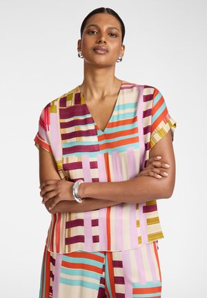 Vrouw met gekruiste armen, draagt een kleurrijke gestreepte V-hals top en bijpassende broek, zilveren oorringen en armband op een witte achtergrond.