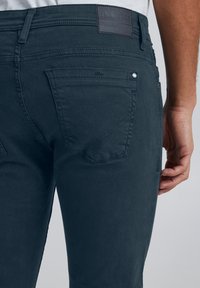 Temno modre kavbojke iz jeansa z oprijetim krojem, zadnjimi žepi z oblikovanimi preklopi in teksturirano rjavo etiketo na pasu.