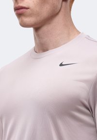 Κοντινό πλάνο ενός ατόμου που φοράει ένα ανοιχτό ροζ πουκάμισο Nike με στρογγυλή λαιμόκοψη και μαύρο λογότυπο Nike στο στήθος.