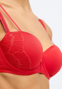 Crvena brassiere sa dvostrukim tankim trakama, glatkim čašama i teksturiranim čipkastim detaljem s jedne strane. Ukrašena malim zlatnim akcentom.