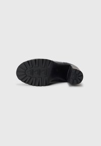 Black shoe sole with ridged texture, featuring triangular traction patterns and a sturdy heel. Minimalistic design and solid construction.