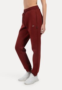 Maronfarvede joggerbukser lavet af blødt stof, med elastisk talje, sidelommer og ribbede manchetter. Sammen med hvide sneakers.
