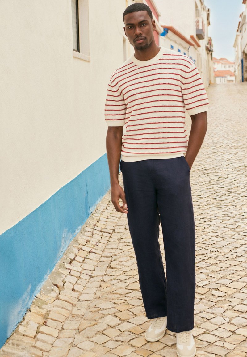 Stribet kortærmet strikket sweater med røde og marineblå striber, parret med marineblå bredbenede bukser og beige sneakers, sat mod en brostensbelagt gade.
