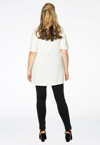 Weiße Kurzarm-Tunika mit längerem Rücken, kombiniert mit schwarzen engen Jeans und Absatzsandalen, von hinten betrachtet.