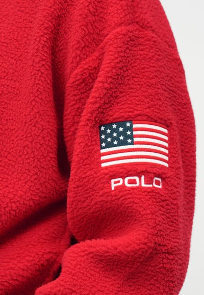 Κόκκινο φλις πουλόβερ με κεντημένη την αμερικανική σημαία και το κείμενο "POLO" στο δεξί μανίκι. Μαλακό, υφασμάτινο υλικό με άνετη εμφάνιση.