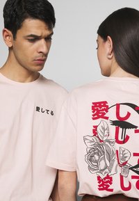 Ljusrosa bomulls-T-shirt med svart japansk text på framsidan och en grafisk design med rosor och detaljer i rött och svart på baksidan.