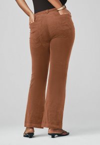 Pantalones acampanados de pana marrón con bolsillos traseros cosidos, que presentan una textura suave y un diseño de cintura alta. Lleva puestos con sandalias de tacón negras.