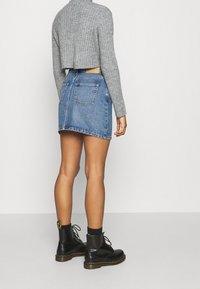 Persona con un suéter gris corto, falda mini de mezclilla azul, calcetas negras y botas de tobillo negras con cordones, de pie contra un fondo plano.