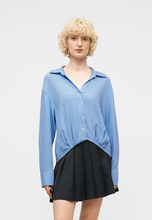 Osoba o krótkich kręconych blond włosach, ubrana w luźną niebieską koszulę na guziki i czarną plisowaną spódnicę, stojąca na jednolitym tle.