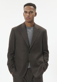 Brown tailored blazer with a notch lapel, two front pockets, and a single button closure, worn over a gray crew-neck sweater.