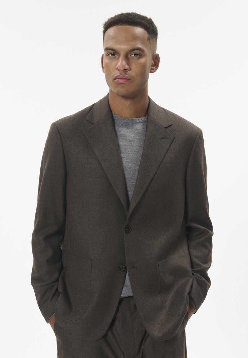 Brown tailored blazer with a notch lapel, two front pockets, and a single button closure, worn over a gray crew-neck sweater.