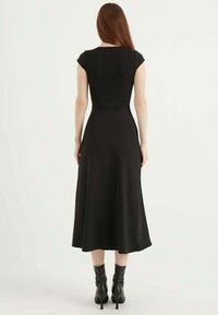 Schwarzes Kleid mit kurzen Ärmeln, einem taillierten Oberteil und einem fließenden A-Linien-Rock; glatter Stoff, knielang, kombiniert mit schwarzen Absatzstiefeletten.