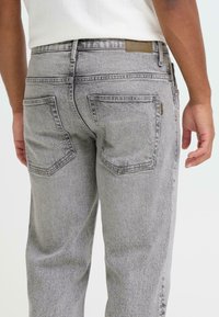 Jean en denim gris à coupe décontractée, avec des poches arrière ornées de détails cousus et une étiquette en cuir sur la ceinture. La texture est douce et usée.