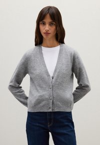Cardigan grigio in morbido tessuto a maglia, con scollo a V, maniche lunghe e chiusura con cinque bottoni. Indossato sopra una camicia bianca con jeans blu.