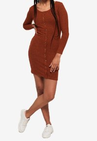 Femme portant une robe boutonnière ajustée à manches longues de couleur rouille avec une texture côtelée, associée à des baskets blanches, se tenant avec une jambe croisée.