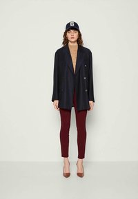 Blazer doppiopetto blu navy sopra un maglione beige a maglia, abbinato a pantaloni aderenti borgogna e tacchi a spillo marroni. Il cappellino presenta il logo "G".