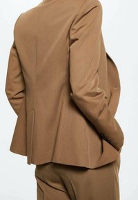 Persona che indossa una giacca di suit marrone su misura e pantaloni abbinati, mostrata di spalle con le mani in tasca su uno sfondo semplice.