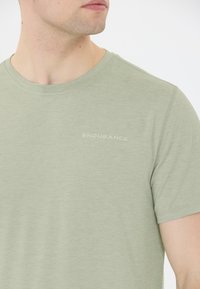 Man som bär en ljusgrön kortärmad skjorta med texten "ENDURANCE" på vänster bröst, tittar åt vänster, mot vit bakgrund.