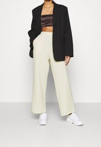 Černý oversize blejzr, hnědý saténový zkrácený top, světle zelené texturované kalhoty širokého střihu a bílé sneakers. Klíčové detaily zahrnují kapsy a texturu.