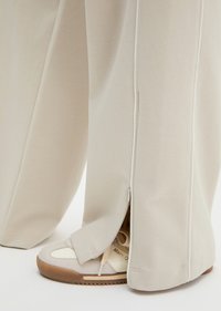 Μπεζ παντελόνια με φαρδιά μπατζάκια με ανοιχτό πλαϊνό φερμουάρ πάνω από λευκά και καφέ αθλητικά παπούτσια σε ανοιχτό φόντο.