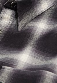 Camicia a quadri grigia e bianca con colletto button-down, caratterizzata da una trama tessuta e bottoni metallici, che mostra un morbido drappeggio del tessuto.