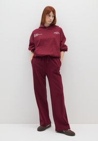 Set di felpa e pantaloni della tuta color bordeaux, con tasca centrale, vita regolabile con cordino e polsini a coste. Il tessuto sembra morbido e strutturato.