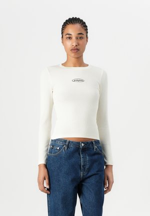 Cremefarbener Langarm-Rippenpullover mit einem Rundhalsausschnitt und einem schwarzen Logo-Patch in der Mitte. Kombiniert mit hochgeschnittenen blauen Jeans.
